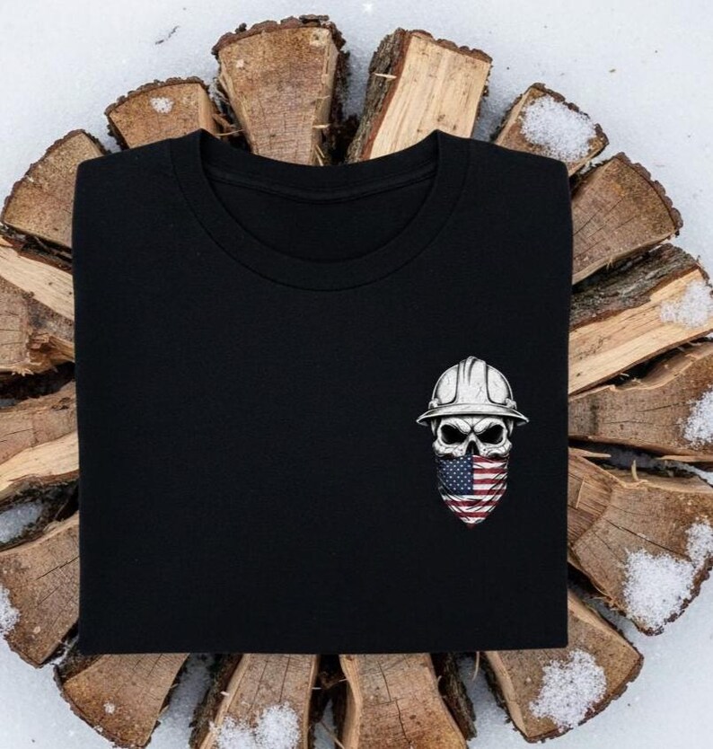 May include: Black t-shirt featuring a skull wearing a white construction helmet and an American flag bandana. The skull design is detailed with shading and highlights. The shirt is displayed on a background of wood and snow.