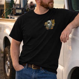 May include: Black t-shirt with a colorful graphic of a welding mask, tools, and the word "WORK" on a flag. The shirt is paired with blue jeans and a brown belt. A black cap is worn.
