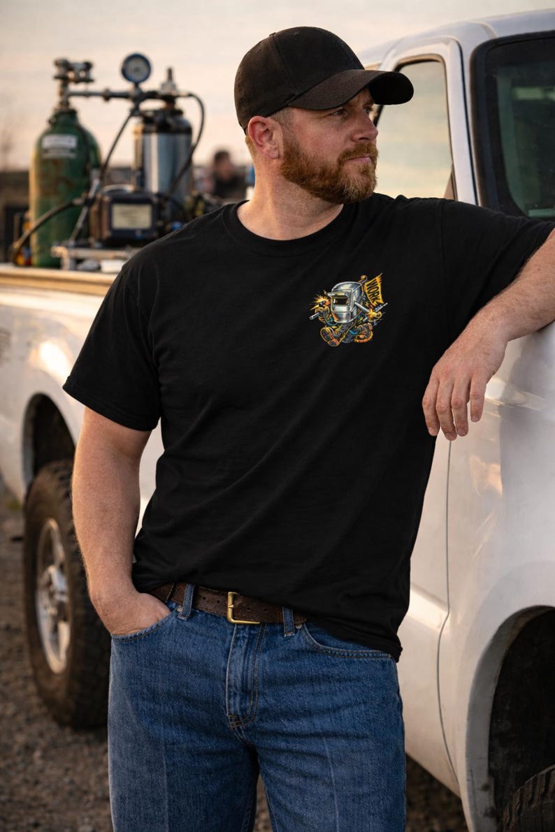 May include: Black t-shirt with a colorful graphic of a welding mask, tools, and the word "WORK" on a flag. The shirt is paired with blue jeans and a brown belt. A black cap is worn.