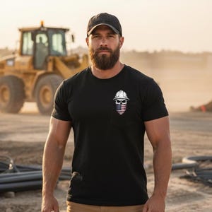 May include: Black crew neck t-shirt with a small pocket featuring a skull wearing a construction helmet and an American flag design. A black baseball cap is worn. A yellow construction vehicle is in the background.