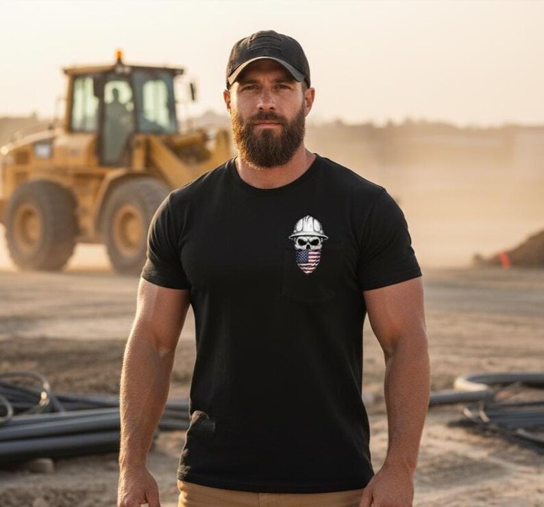 May include: Black crew neck t-shirt with a small pocket featuring a skull wearing a construction helmet and an American flag design. A black baseball cap is worn. A yellow construction vehicle is in the background.