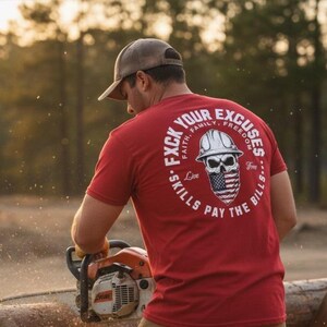 May include: A red t-shirt with a graphic on the back featuring a skull wearing a hard hat and an American flag bandana. The text reads "F*ck Your Excuses, Skills Pay The Bills". The person is using a chainsaw.