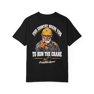 Może przedstawiać: Czarny t-shirt z grafiką brodatego pracownika budowlanego w żółtym kasku z amerykańską flagą, wskazującego palcem przed siebie. Napis głosi THIS COUNTRY NEEDS YOU TO RUN THE CRANE oraz Craneaholics Apparel z ikoną dźwigu.