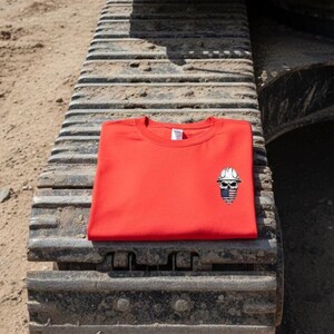 May include: A bright red crew neck sweatshirt with a small embroidered design of a skull wearing a construction hard hat. The skull has a blue and white striped design. The sweatshirt is folded and resting on a construction vehicle track.