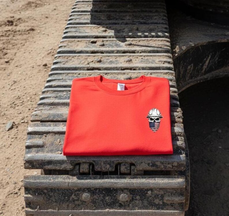May include: A bright red crew neck sweatshirt with a small embroidered design of a skull wearing a construction hard hat. The skull has a blue and white striped design. The sweatshirt is folded and resting on a construction vehicle track.