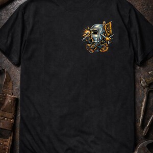 May include: Black t-shirt featuring a cartoon welder with a welding mask, holding a torch, and a flag that says "WORK". The graphic is in shades of gray, orange, and yellow. The shirt is a crew neck.