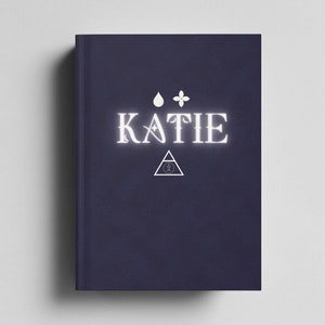 May include: A dark blue book cover with the name "KATIE" in white text. The cover also features a white teardrop, a white flower, and a white triangle with a circle inside.