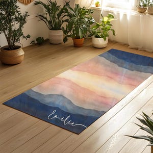 May include: A personalized yoga mat with a watercolor design of mountains and a sunset. The mat is blue, pink, and yellow with the name "Emilie" written in white cursive.