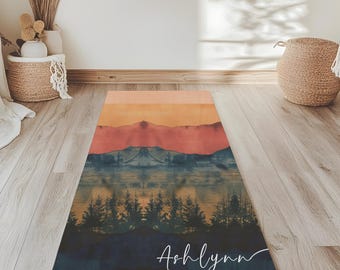 Puede incluir: Una esterilla de yoga personalizada con un diseño de paisaje de montaña al atardecer. La esterilla es de color azul oscuro con una escena de montaña azul claro, naranja y rojo. El nombre "Ashlynn" está escrito en cursiva blanca en la parte inferior de la esterilla.