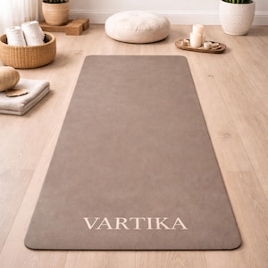Puede incluir: Una esterilla de yoga rectangular de color topo neutro, con la palabra "VARTIKA" impresa en blanco en la parte inferior. La esterilla está colocada sobre un suelo de madera clara, con una cesta de toallas enrolladas y un cojín pequeño. Medidas en cm.