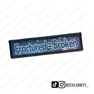 May include: A rectangular patch with the words "Fractured = Broken" in white, outlined in light blue. The background is a dark blue with colorful splatters. The patch has a black border.
