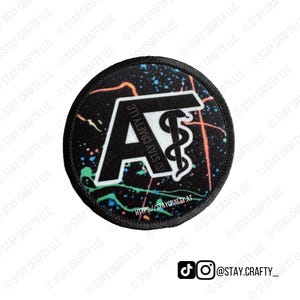 May include: A round black patch with a white "A" and a medical symbol. The patch has a colorful splatter design in orange, blue, and green. The text "STAY CRAFTY LLC" is printed around the edge and on the design.
