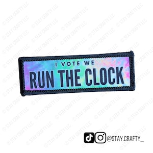 May include: A rectangular patch with the text "I VOTE WE RUN THE CLOCK" in black letters. The background has a tie-dye effect with shades of blue, purple, and green. The patch is bordered with black stitching.