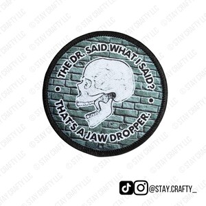 May include: Round patch with a black border featuring a skull illustration and the text "THE DR. SAID WHAT I SAID? THAT'S A JAW DROPPER." The background is a brick wall design. The patch also includes social media handles.
