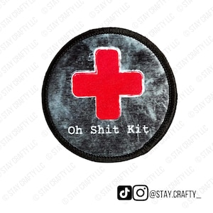 May include: Round patch with a red cross on a gray background, encircled by a black border. The text "Oh Shit Kit" is displayed below the cross. The patch has a distressed, vintage look.