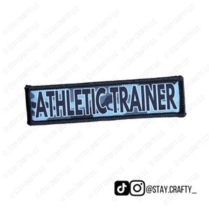 May include: A rectangular patch with the words "ATHLETIC TRAINER" in white block letters. The background has a blue camouflage pattern. The patch is bordered with black stitching.