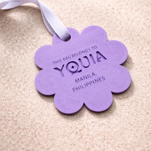 May include: A lavender, flower-shaped tag with the text "THIS BAG BELONGS TO YOUIA, MANILA, PHILIPPINES." The tag is attached to a white ribbon. The background is a light tan color.