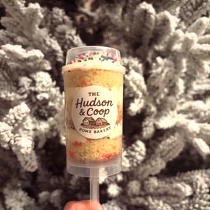 May include: A push-pop cake with layers of yellow cake and frosting, topped with colorful sprinkles. The clear plastic container features a label that reads "The Hudson & Coop Home Bakery."