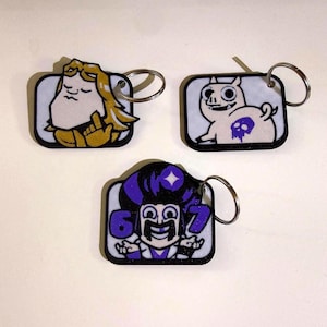 May include: Three cartoon character keychains. One features a figure with blonde hair and a gold outfit, another a pig with a skull, and the last a character with purple hair and the numbers 6 and 7. Each keychain has a silver ring.