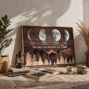 May include: A framed wooden print featuring four moon phases with names and dates. The text "The days you became our Dad" is below the phases. A mountain and forest silhouette is at the bottom. A lantern, books, and a mug are in the foreground.