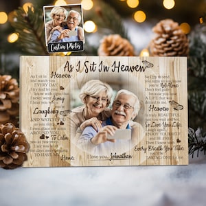 May include: A wooden sign with a heart shape in the center. The sign has a light wood background with a darker wood grain pattern. The sign features a photo of an older couple smiling and taking a selfie. The text on the sign reads "As I Sit In Heaven" and includes a poem about love, loss, and remembering a loved one.