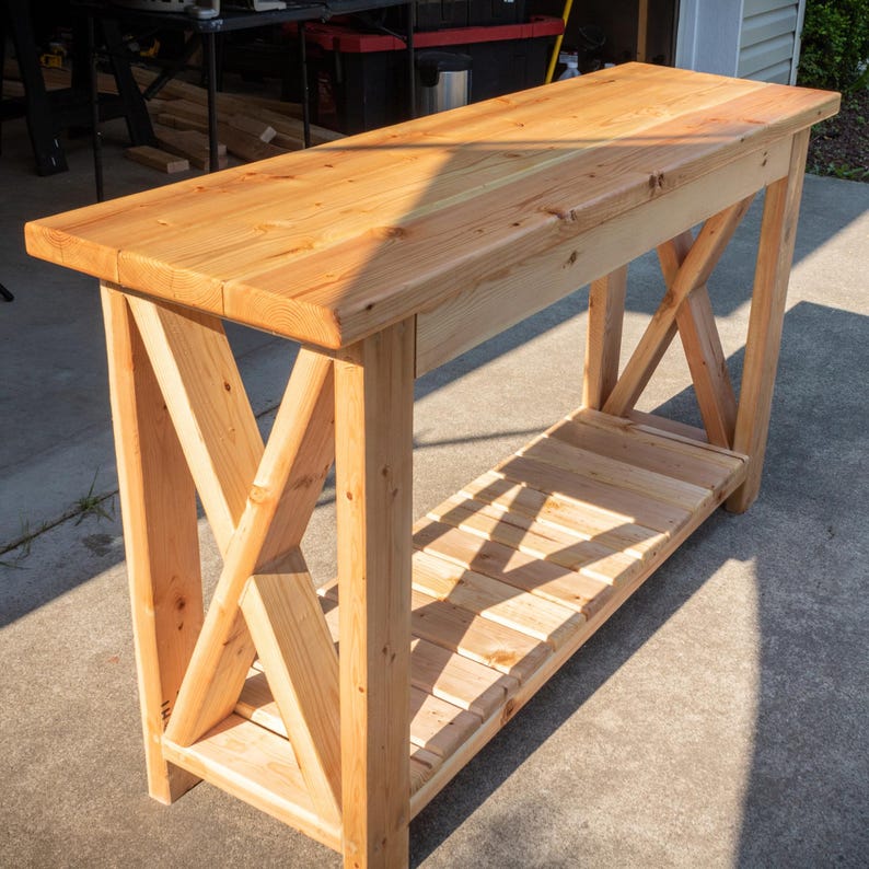 May include: A light-colored wooden console table with an X-shaped support design. The table has a rectangular top and a lower shelf made of wooden slats. The table is constructed from light-colored wood and is suitable for a hallway or living room.