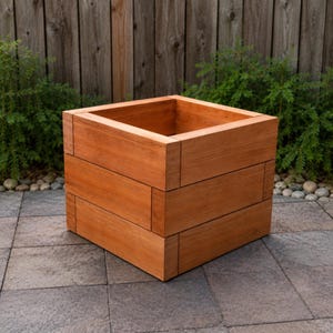 Può includere: Una fioriera quadrata costruita con assi di legno orizzontali con una finitura naturale marrone rossastro. La fioriera presenta un design impilato con giunture visibili dove le assi si incontrano agli angoli e lungo i lati della struttura.