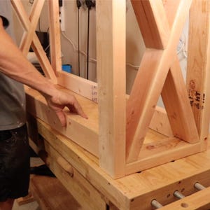 May include: A partially constructed wooden table frame with an X-shaped support design. The natural wood has a light brown color. The table is in the process of being built, with visible joinery and unfinished surfaces.