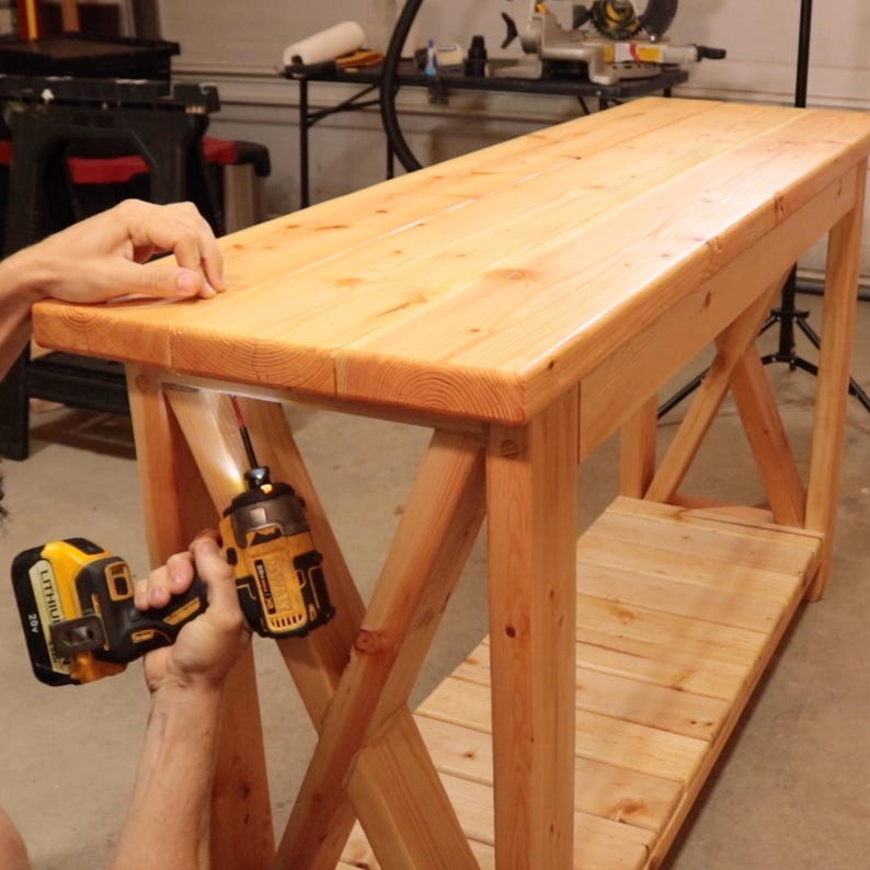 May include: A light-colored wooden console table with an X-shaped support design. The table is being assembled with a yellow and black power drill. The table has a lower shelf for storage and is made of natural wood.