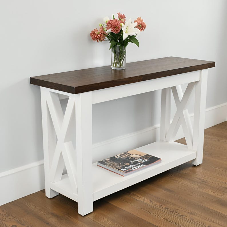 May include: A white and brown console table with an X-frame design. The table has a dark brown top and a lower shelf. A clear glass vase with flowers sits on top of the table. A magazine rests on the shelf.
