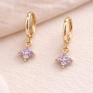 May include: A pair of gold-colored hoop earrings, each with a dangling charm. The charms are four-petal flower shapes, each petal set with a light purple gemstone. The earrings are displayed on a cream-colored fabric.
