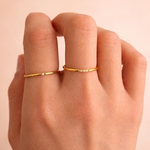 May include: Two gold-colored rings with small, clear gemstones. One ring has a single gemstone, while the other has three. The rings are thin and delicate, and are displayed on a hand against a neutral background.