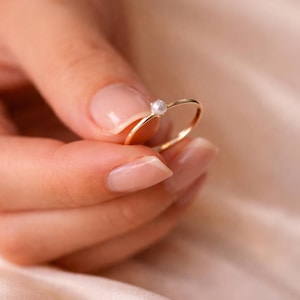 May include: A thin gold ring featuring a single small, round white pearl set at the top. The band is smooth and circular, showcasing a polished metallic finish that contrasts with the delicate, lustrous appearance of the centered pearl gemstone.