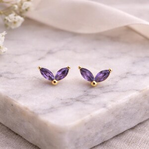 May include: A pair of gold-colored stud earrings with a leaf-like design. Each earring features three purple marquise-cut gemstones and a small gold ball. The earrings are displayed on a marble surface.