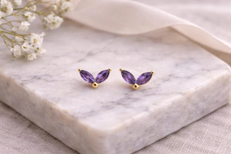 May include: A pair of gold-colored stud earrings with a leaf-like design. Each earring features three purple marquise-cut gemstones and a small gold ball. The earrings are displayed on a marble surface.
