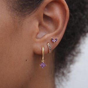 May include: A close-up of an ear adorned with three gold earrings. One is a small stud in the shape of a butterfly, another is a curved bar with purple stones, and the third is a hoop with a dangling square purple stone.