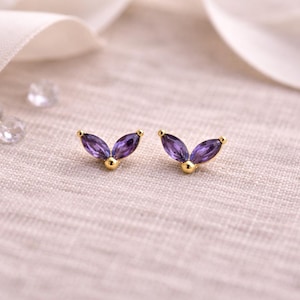May include: A pair of gold-colored stud earrings with a leaf-like design. Each earring features two marquise-cut, deep purple gemstones and a small gold ball at the base. The earrings are set against a soft, textured background.