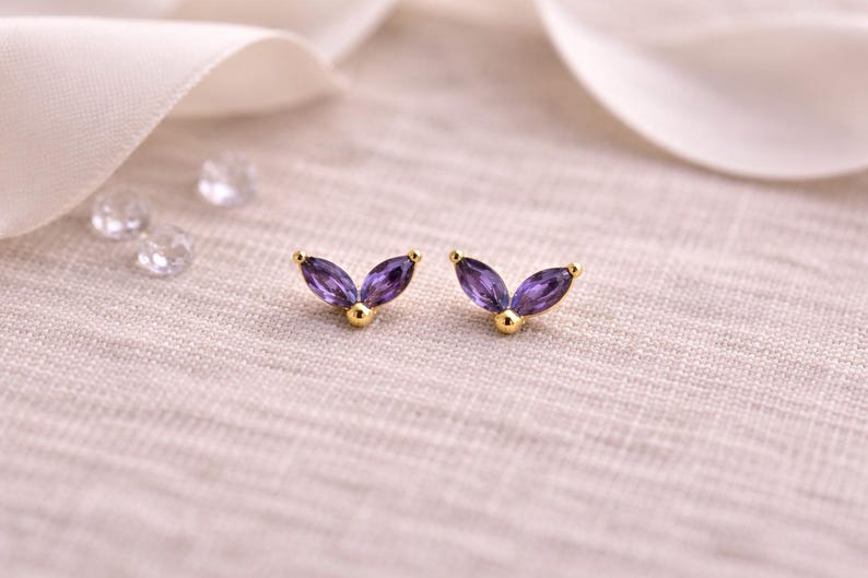 May include: A pair of gold-colored stud earrings with a leaf-like design. Each earring features two marquise-cut, deep purple gemstones and a small gold ball at the base. The earrings are set against a soft, textured background.