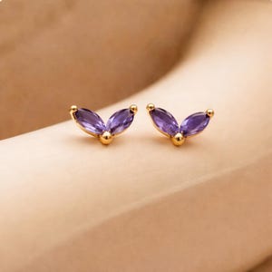 May include: A pair of gold stud earrings, each featuring a marquise-cut, deep purple gemstone. The earrings have a delicate, leaf-like design with a small gold ball at the center and at the end of each gemstone.