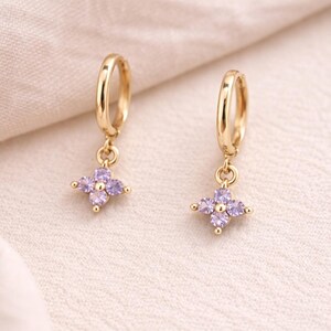 May include: A pair of gold-colored hoop earrings. Each earring features a small, dangling charm in the shape of a four-petal flower, adorned with light purple gemstones. The earrings are displayed on a cream-colored fabric.