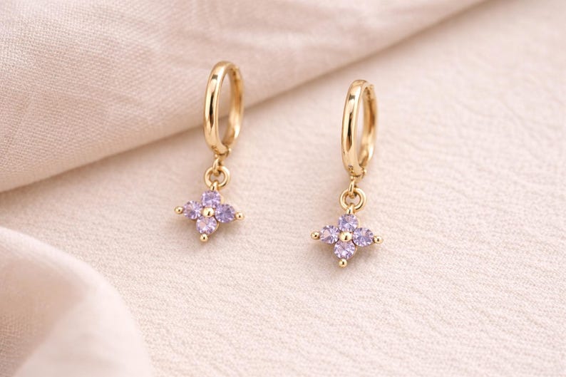 May include: A pair of gold-colored hoop earrings. Each earring features a small, dangling charm in the shape of a four-petal flower, adorned with light purple gemstones. The earrings are displayed on a cream-colored fabric.