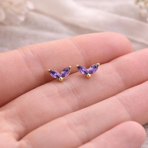 May include: A pair of gold-colored stud earrings, each featuring three marquise-cut purple gemstones arranged in a V-shape. The earrings are displayed on a light-colored background.