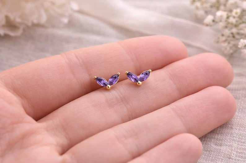 May include: A pair of gold-colored stud earrings, each featuring three marquise-cut purple gemstones arranged in a V-shape. The earrings are displayed on a light-colored background.