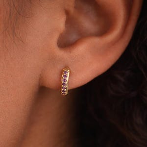May include: A gold-colored hoop earring with a row of small, purple gemstones. The earring is worn on an ear, with the skin tone appearing warm and natural. The earring's design is simple and elegant, suitable for everyday wear.
