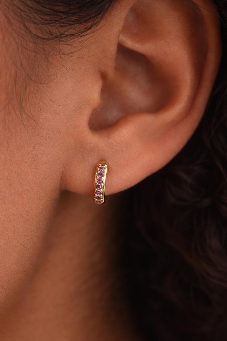 May include: A gold-colored hoop earring with a row of small, purple gemstones. The earring is worn on an ear, with the skin tone appearing warm and natural. The earring's design is simple and elegant, suitable for everyday wear.