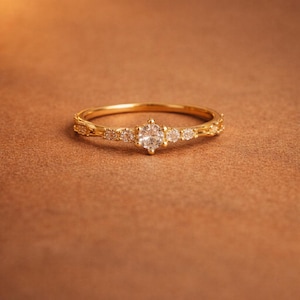 May include: A gold ring with a central, round, clear gemstone and smaller clear gemstones on either side. The band is gold and has a delicate, textured design. The ring is set against a warm, brown background.