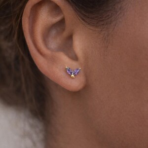 May include: A close-up of a person's ear with a gold stud earring. The earring features two purple marquise-cut gemstones and a small gold ball. The earring is set in gold and is a delicate piece of jewelry.