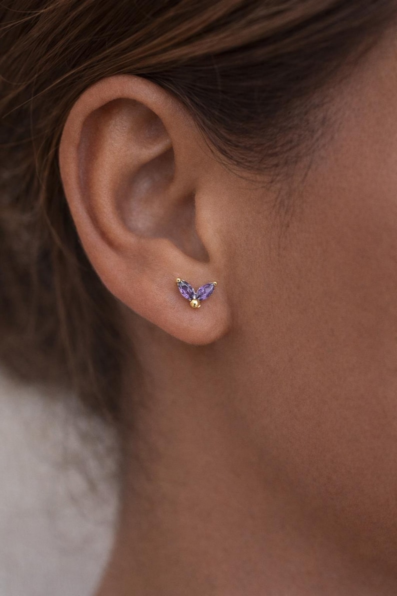 May include: A close-up of a person's ear with a gold stud earring. The earring features two purple marquise-cut gemstones and a small gold ball. The earring is set in gold and is a delicate piece of jewelry.