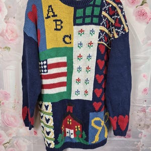 May include: A long-sleeved knitted jumper featuring a patchwork design with various motifs including the letters ABC, an American flag, a red schoolhouse, red hearts, a yellow bow, and floral patterns in colours of navy blue, yellow, red, and green.