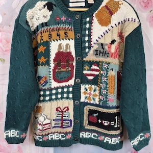 Könnte beinhalten: Eine dunkelgrüne Strickjacke mit einem mehrfarbigen Patchwork-Design und verschiedenen Motiven zum Thema Schule, darunter ein Schaf, ein Rucksack, ein Bleistift, ein Milchkarton, Bücher und die Buchstaben ABC. Die Jacke hat eine Knopfleiste.